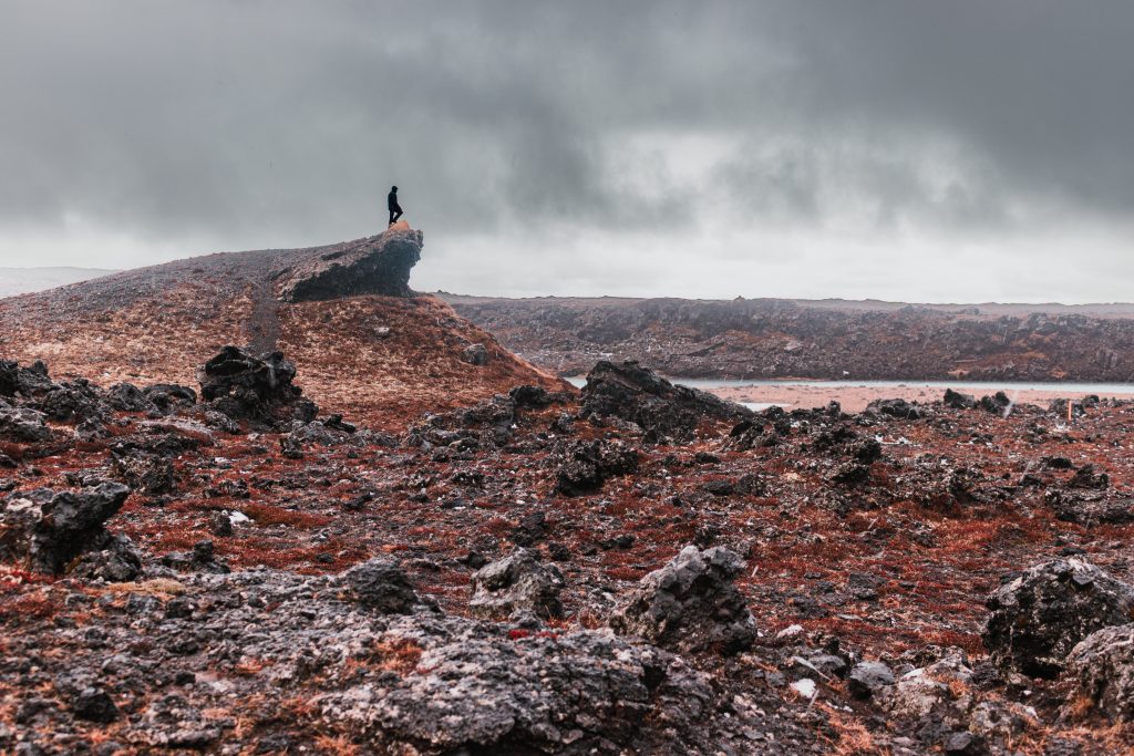 Landscape photograph of a lone silhouette standing atop a jagged volcanic crater ridge in Iceland under an overcast sky